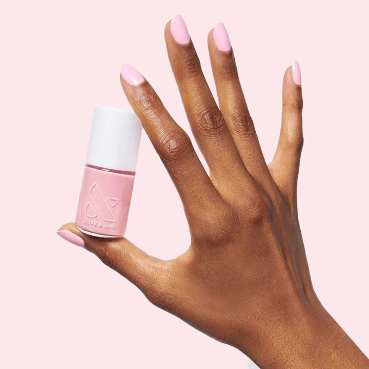 Hand with nails painted soft pink holding SE nail polish bottle against pale pink background for mani look