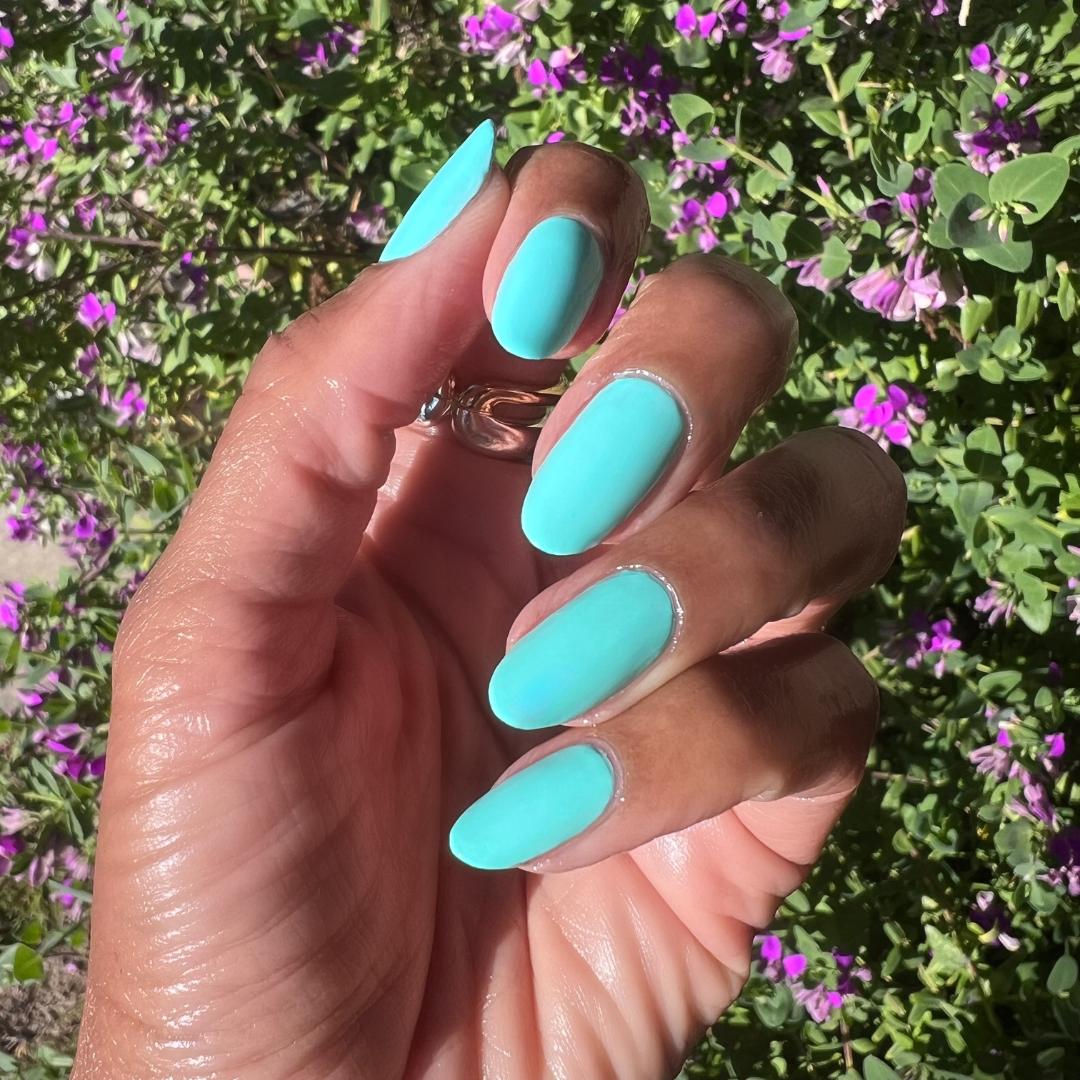 Hand with nails painted in bright blue-green All Manis on Deck gel polish showing smooth manicure outdoors with floral background