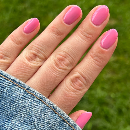 Hand with nails painted in bright pink Taffy gel polish shown outdoors with denim jacket sleeve and green grass background