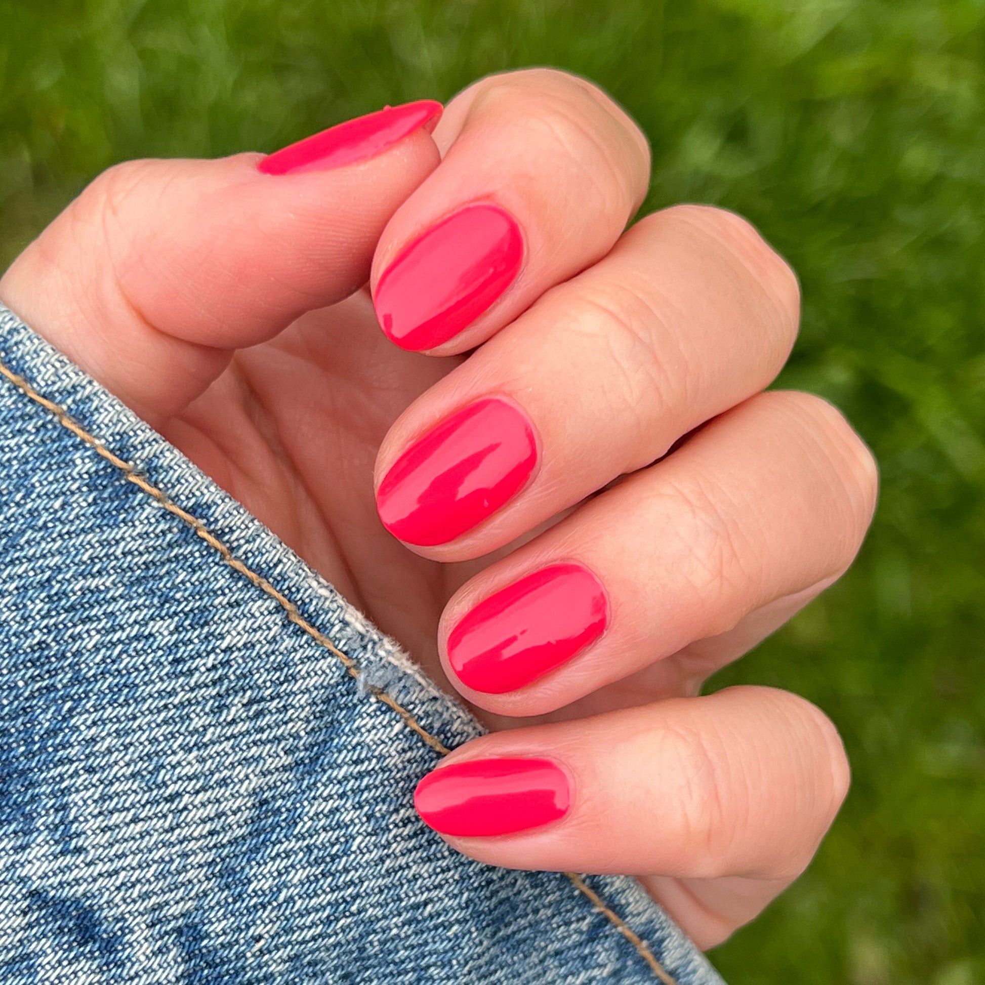 Close-up of hand with Pomodoro quick dry polish in glossy pinky-tomato red on natural nails near denim fabric outdoors