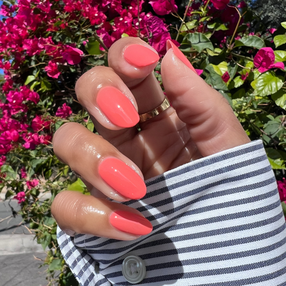 Hand with almond-shaped nails painted in Cape showing a glossy pinky-red quick dry manicure outdoors with bright flowers background