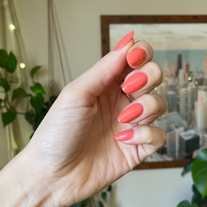Hand with oval nails painted in Cape showing a glossy preppy pinky-red manicure with a city skyline blurred background