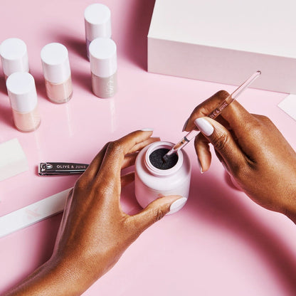 Hands holding the Clean Up Brush dipping into black polish with nail care tools and polish bottles on pink background
