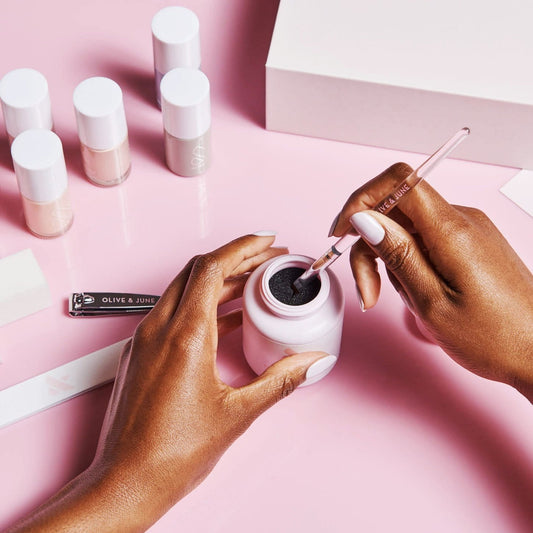 Hands holding the Clean Up Brush dipping into black polish with nail care tools and polish bottles on pink background