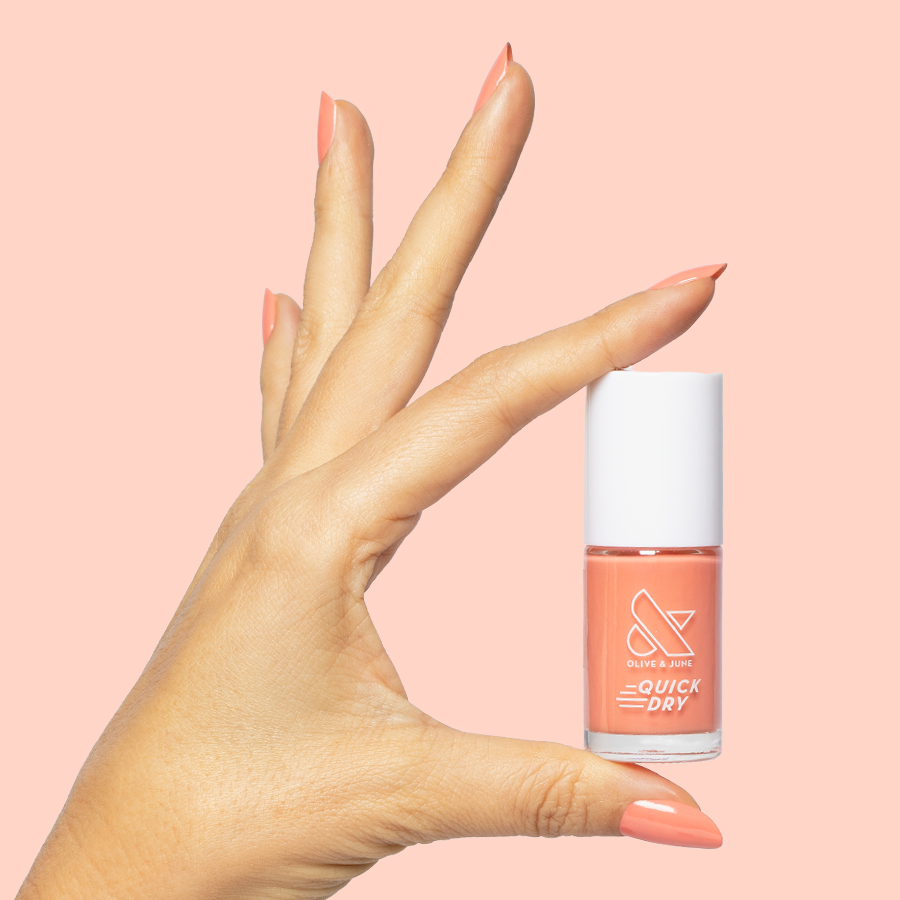 Hand holding Frolic bright orangey coral quick dry gel polish bottle with matching mani on soft peach background