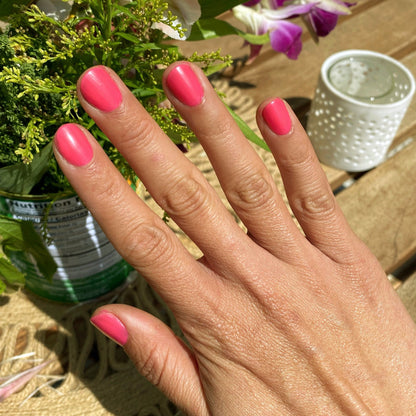Hand with nails painted in vibrant Hot Strawberry gel polish in bright strawberry pink next to flowers on wooden surface