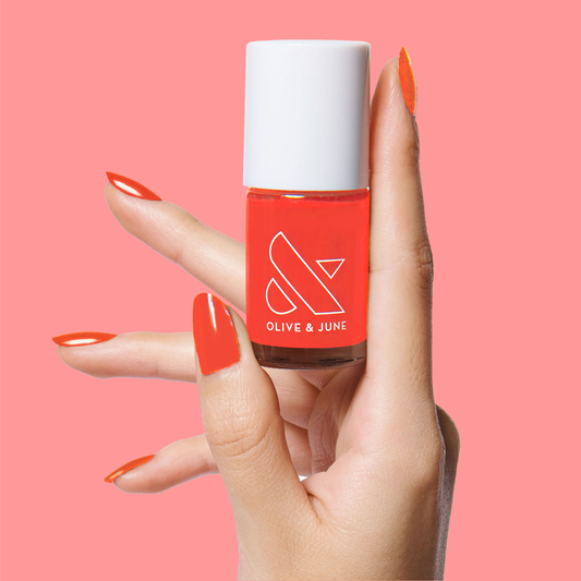 Hand holding Olive and June Lava nail polish with matching bright spicy orange-red mani on coral background