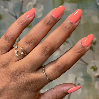 Hand with glossy almond-shaped nails painted in bright orangey coral Frolic gel polish with delicate gold rings on neutral floral background