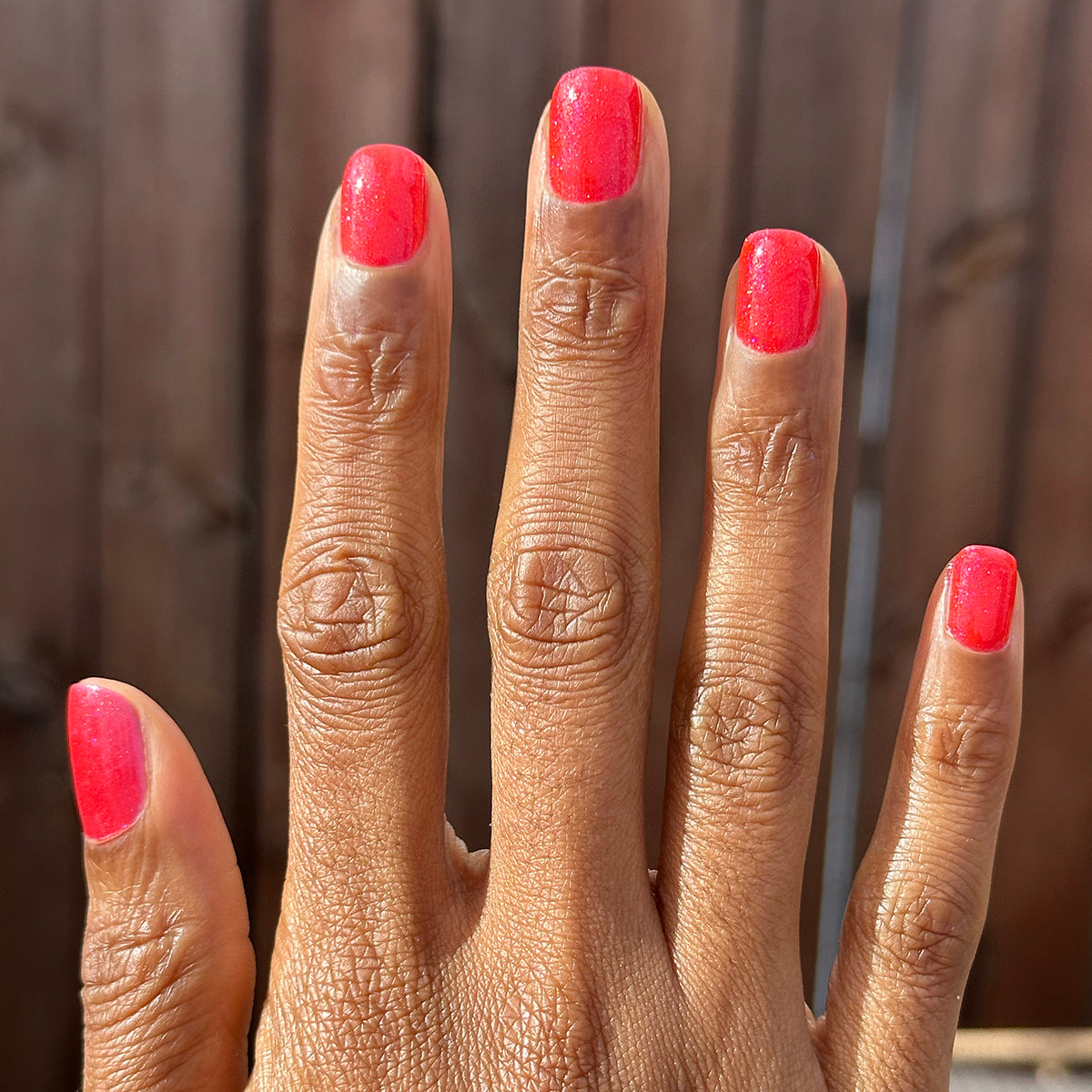 Hand with medium-length nails painted bright pinky-red shimmer using Rodeo Queen - Gel polish in natural light