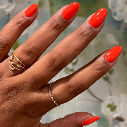 Hand with medium-length nails painted in bright Snapdragon orange-red gel polish with gold and silver rings on floral background